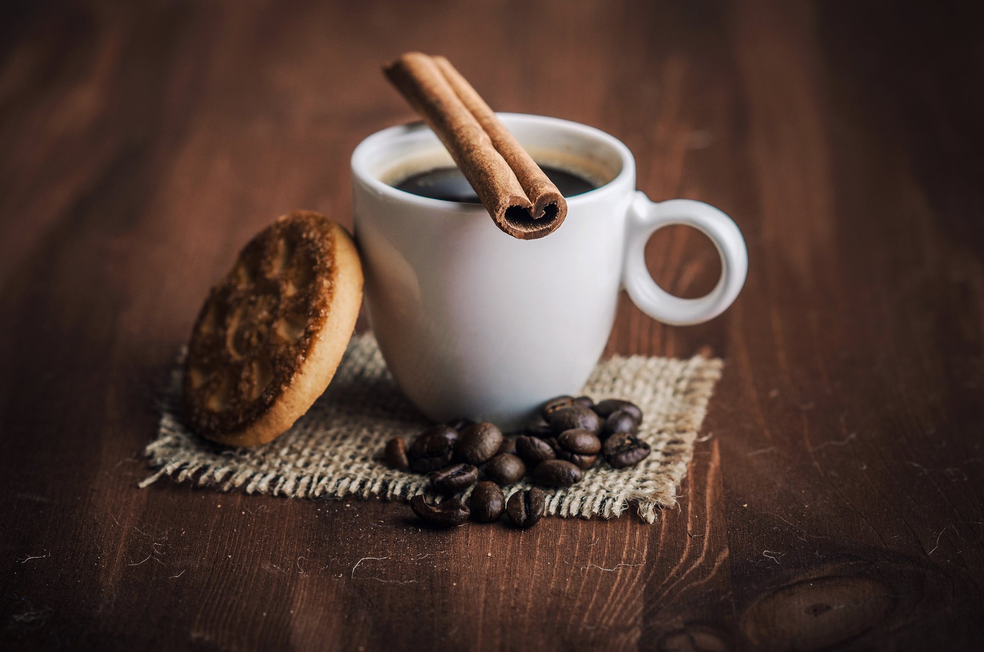 На деревянном столе белая чашка с кофе, корицей, печененкой и кофейными зёрнами