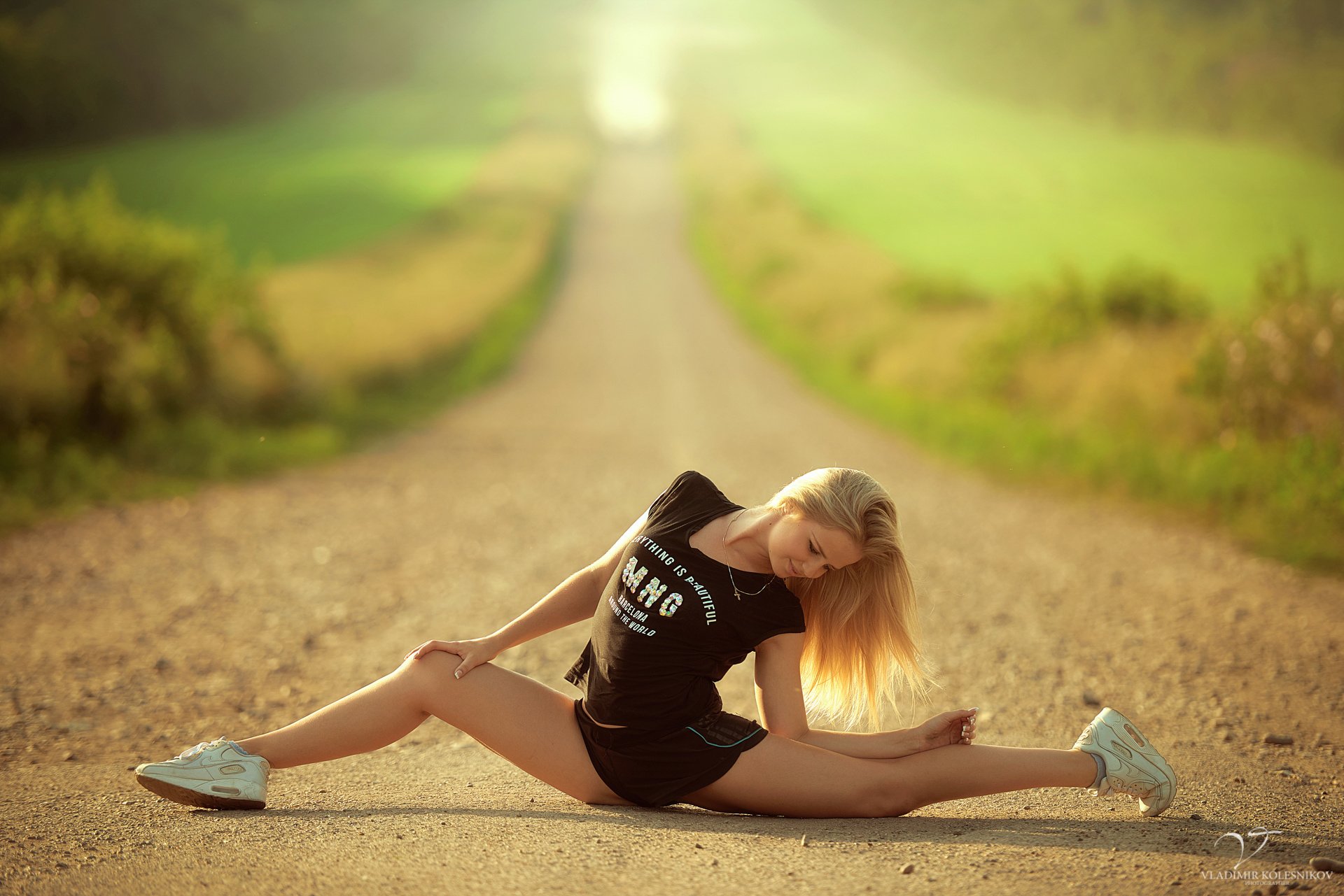 стройная спортивная девушка блондинка села посреди дороги поза растяжка шпагат гибкость футболка шортики кроссовки солнце зелень размытие